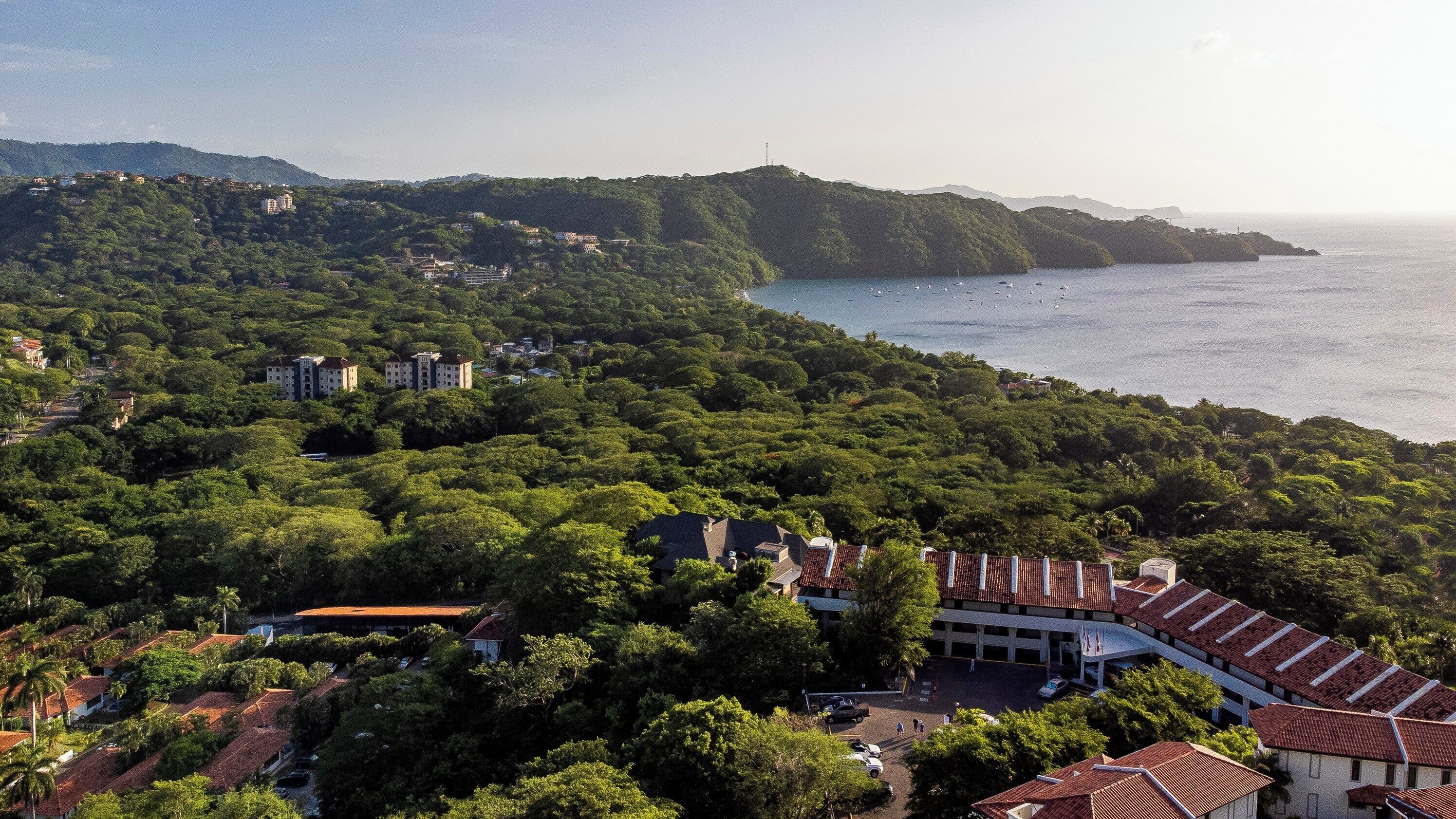 A coastal area with dense greenery, buildings with red-tiled roofs, and a view of the sea with boats.