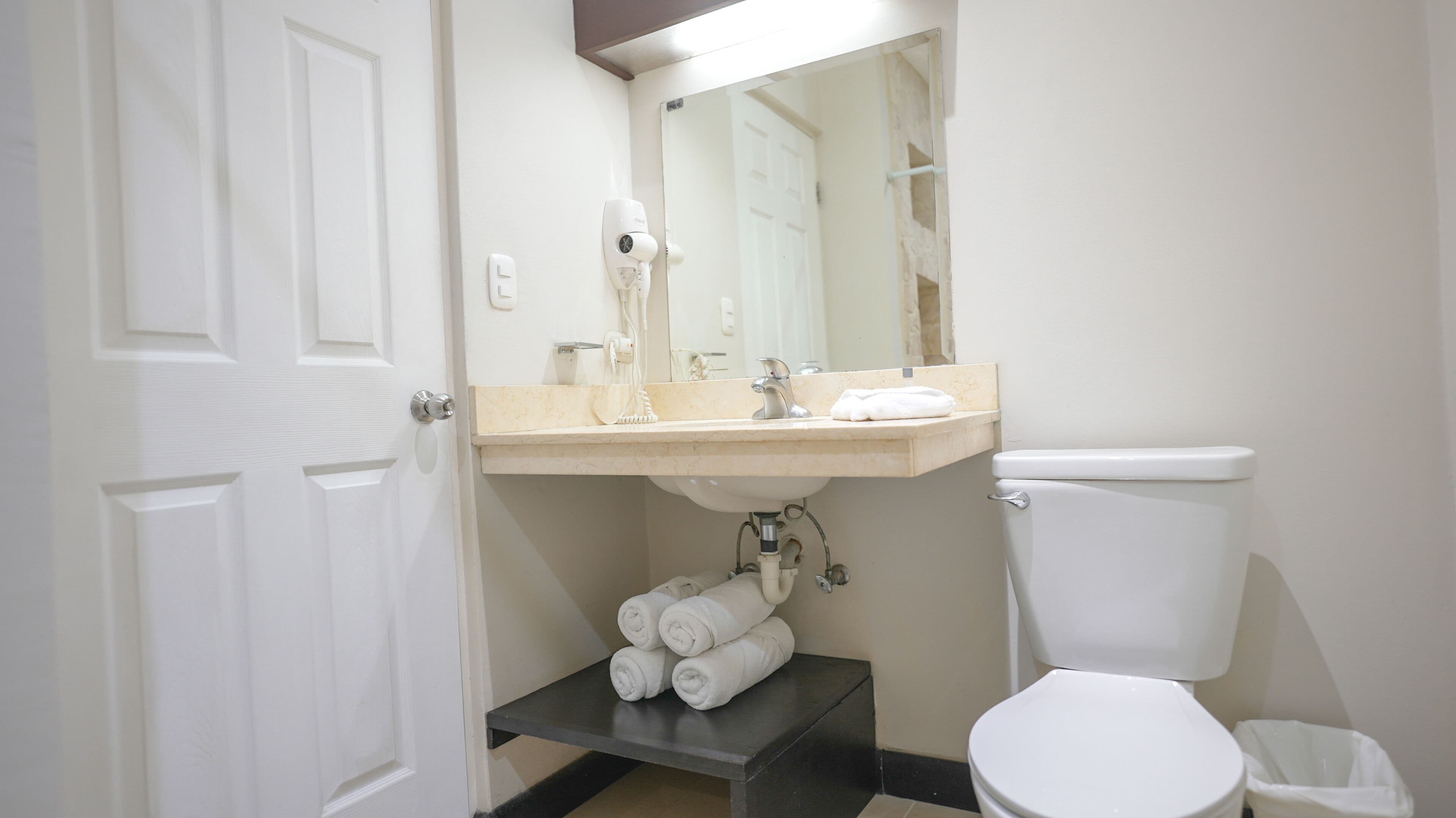 Un baño con inodoro blanco, un lavabo con espejo y un secador de pelo montado en la pared. Hay toallas apiladas en un estante debajo del lavabo.