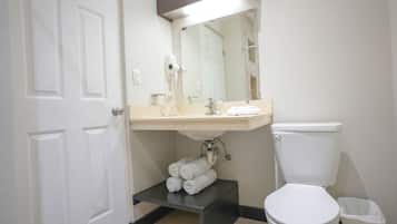 Un baño con inodoro blanco, un lavabo con espejo y un secador de pelo montado en la pared. Hay toallas apiladas en un estante debajo del lavabo.