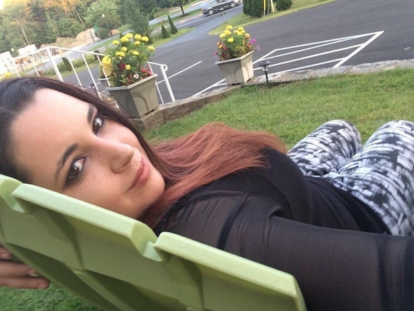 A person reclining in a green chair outdoors with a road and flower pots in the background. - Robin Hood Motel (Ballston Spa)