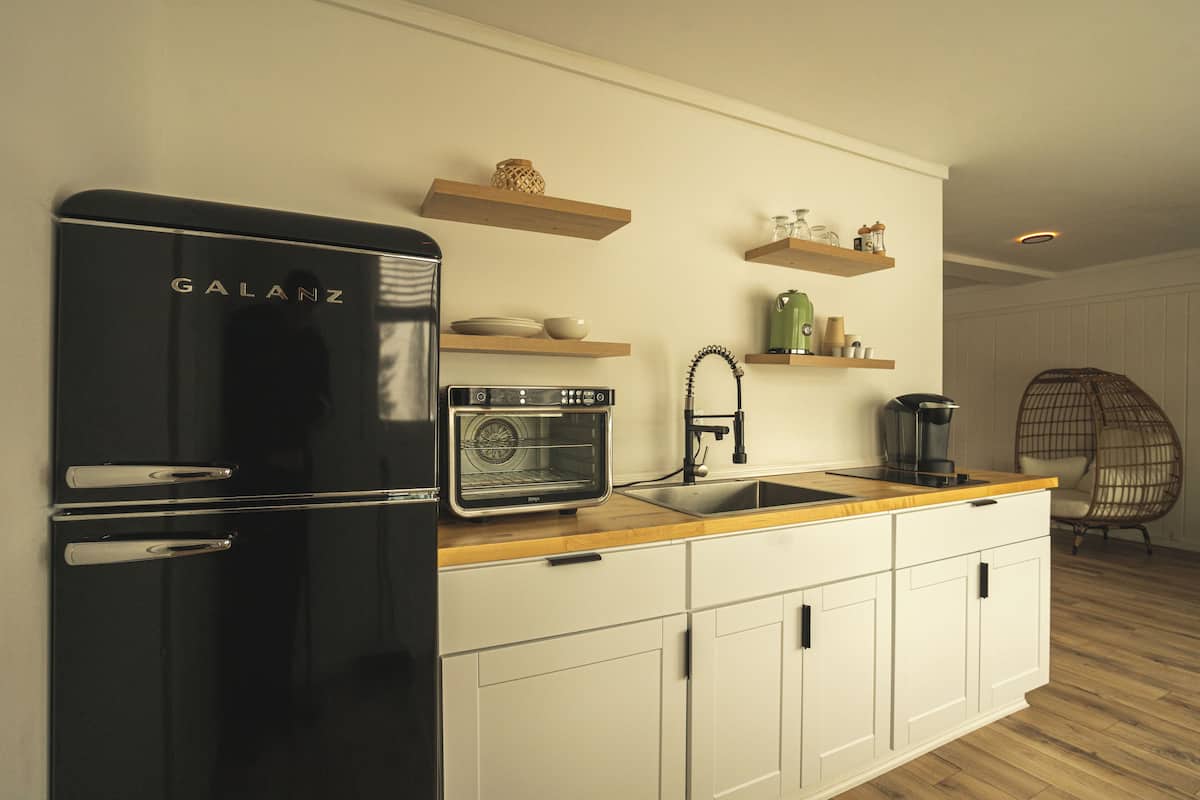 A modern kitchen with a black refrigerator, a built-in oven, a sink, wooden shelves, and a wicker chair.