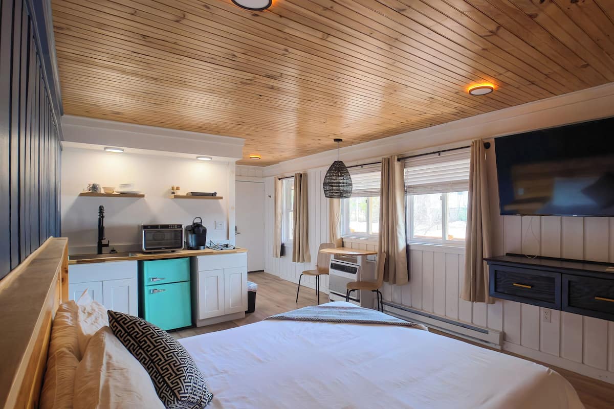 A bedroom with a wooden ceiling, a bed, a TV, a kitchenette, and a window with curtains.