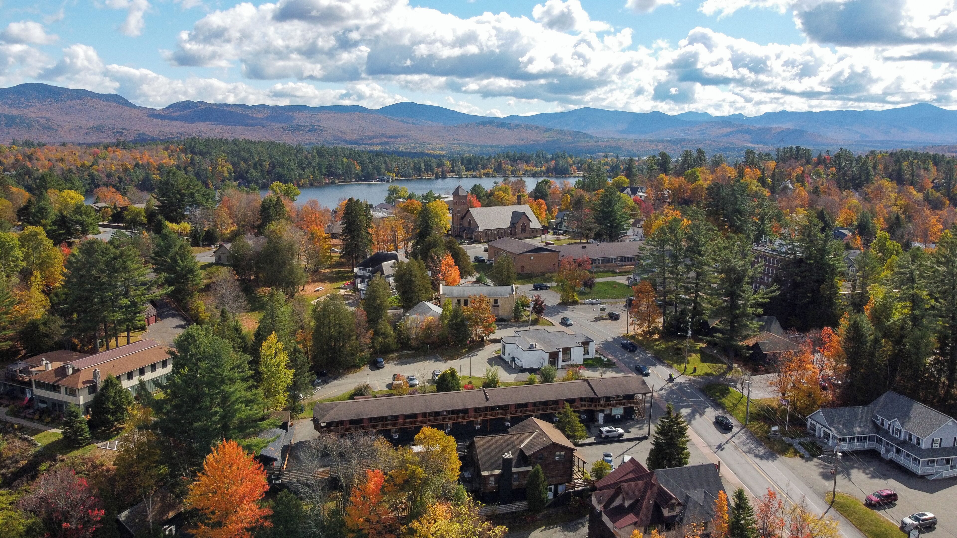 An aerial view of a residential area with houses, a church, and a lake surrounded by mountains.