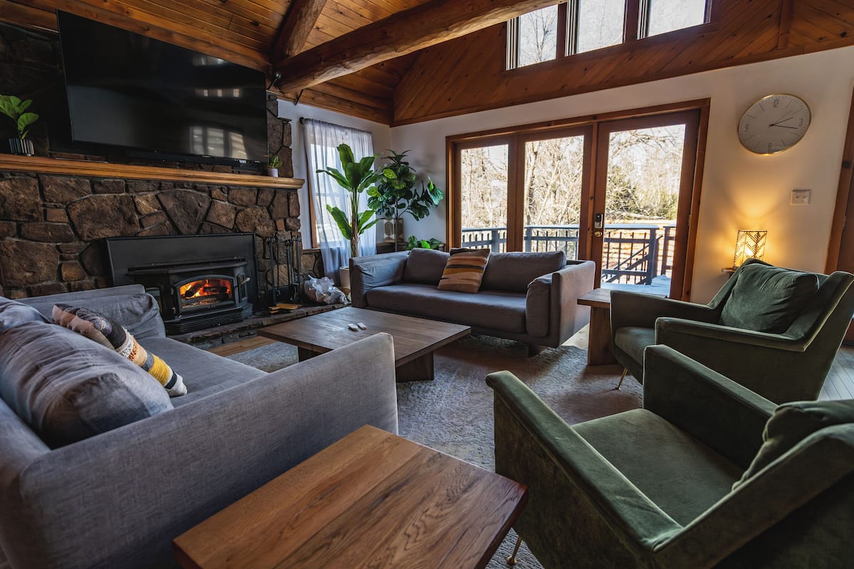 A cozy living room with a stone fireplace, comfortable seating, and a large window offering a view of the outdoors.