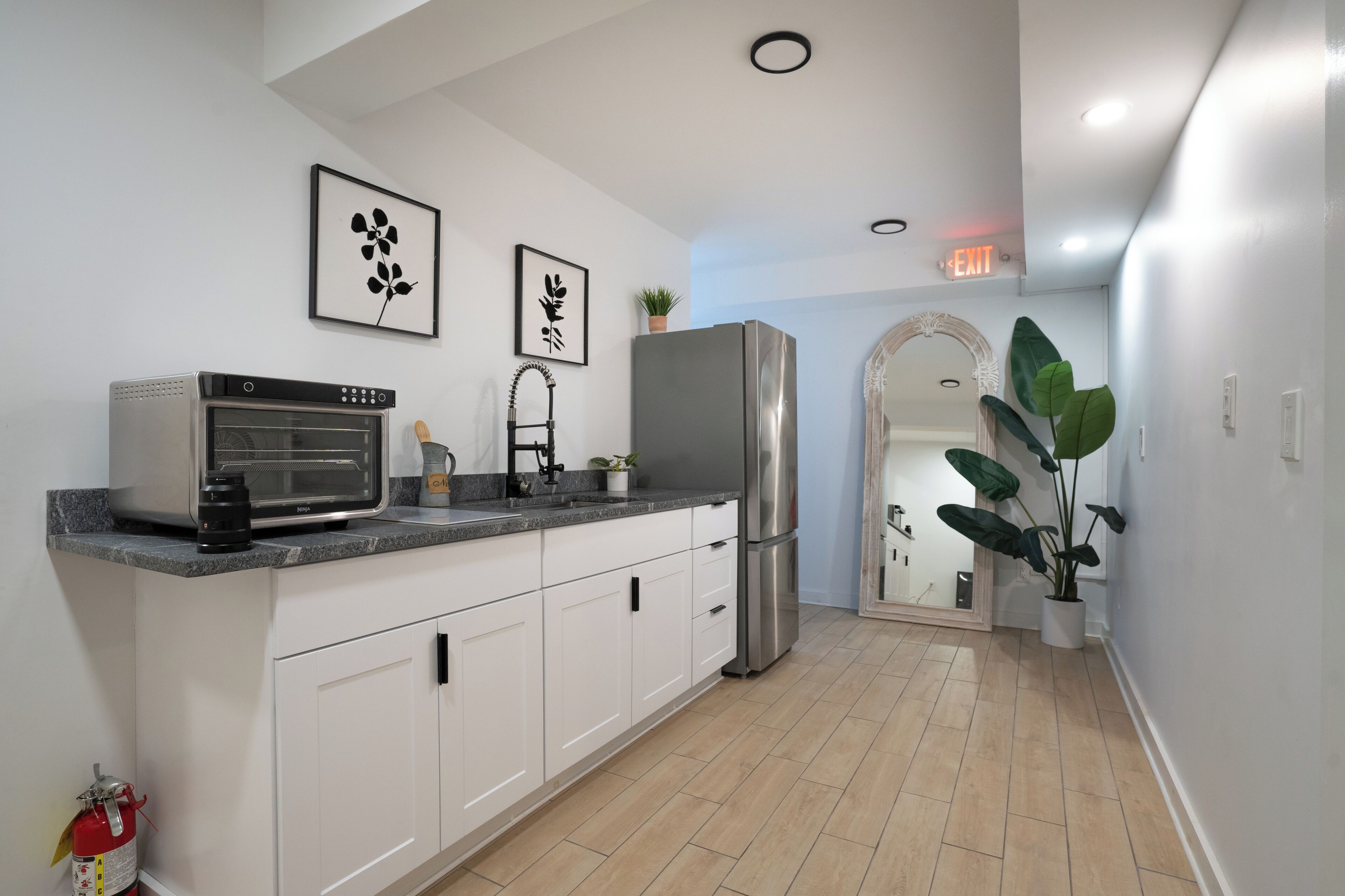 A modern kitchen with white cabinets, a stainless steel refrigerator, a built-in oven, and a microwave. There are two framed wall art pieces, a potted plant, and a fire extinguisher on the counter.