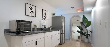 A modern kitchen with white cabinets, a stainless steel refrigerator, a built-in oven, and a microwave. There are two framed wall art pieces, a potted plant, and a fire extinguisher on the counter.
