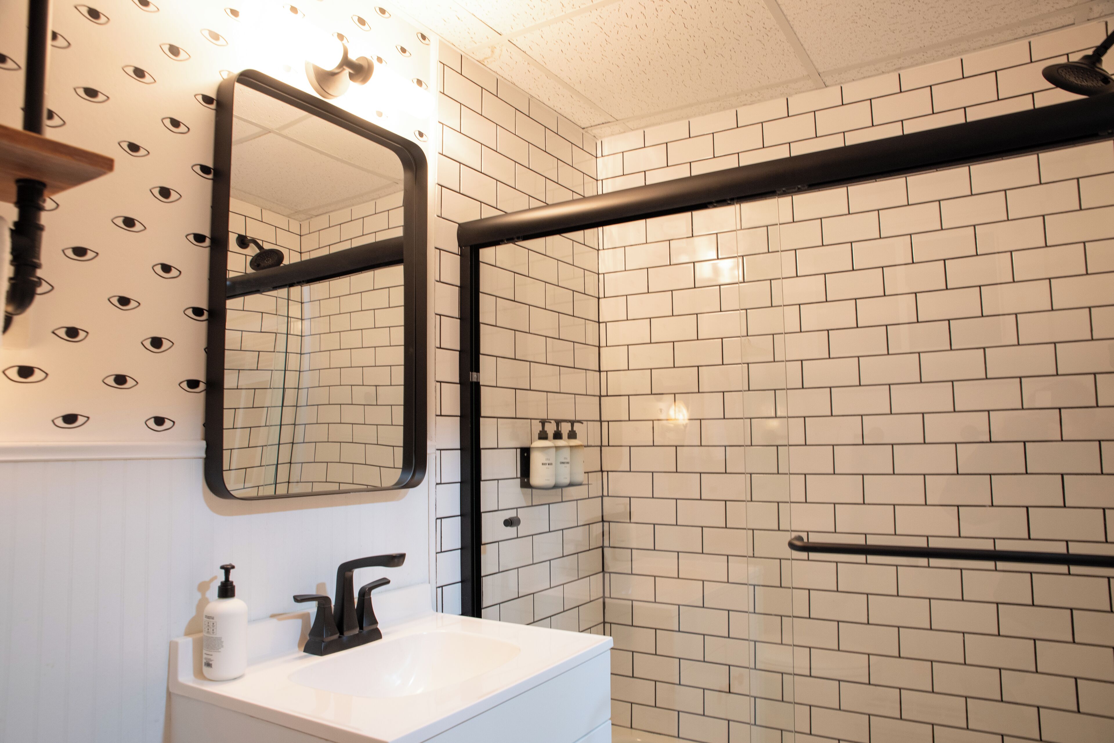 A bathroom with a white sink, black faucet, and a mirror with a black frame. There are white tiles on the walls and a black towel rack.