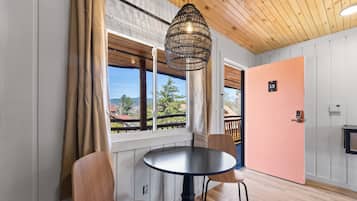 Una habitación con una mesa redonda, dos sillas, una lámpara colgante, una puerta con un número y una ventana con cortinas.