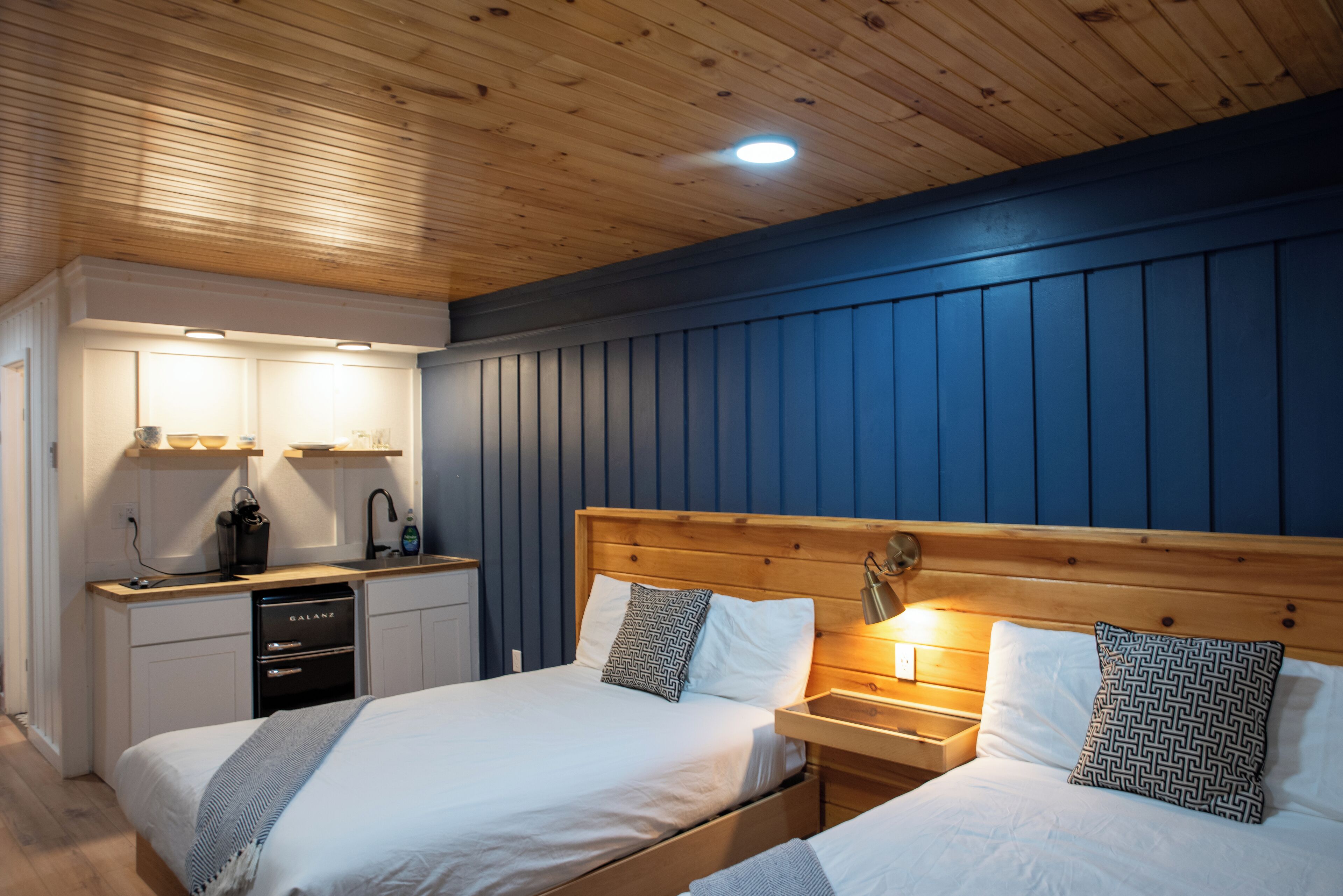 Une chambre avec deux lits, un plafond en bois, un mur bleu, une petite kitchenette et une lampe de chevet.