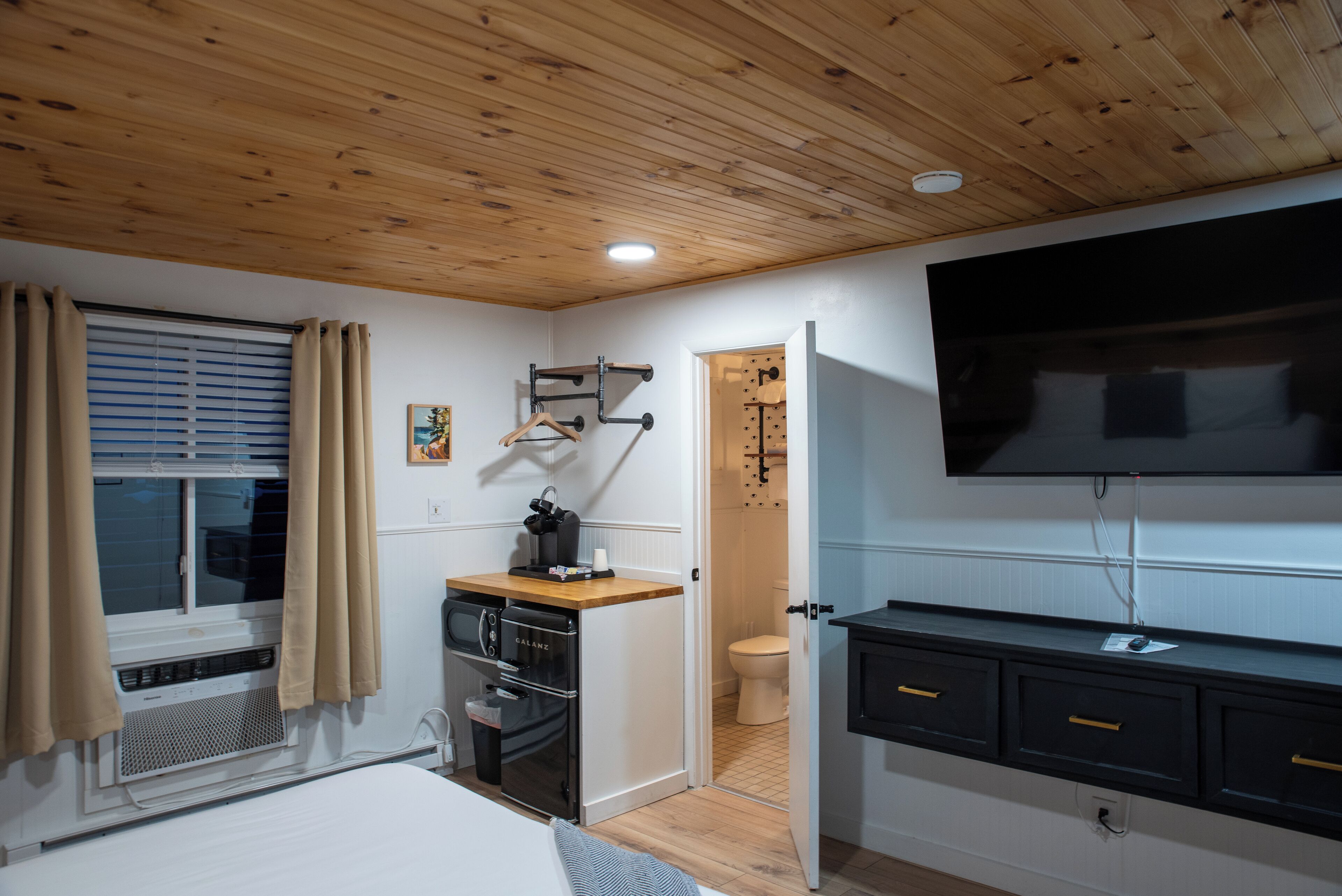 Ein gemütlicher Raum mit Holzbalkendecke, einem Bett, einer kleinen Küchenzeile mit Herd, einem an der Wand montierten Fernseher und einer Tür zu einem Badezimmer.