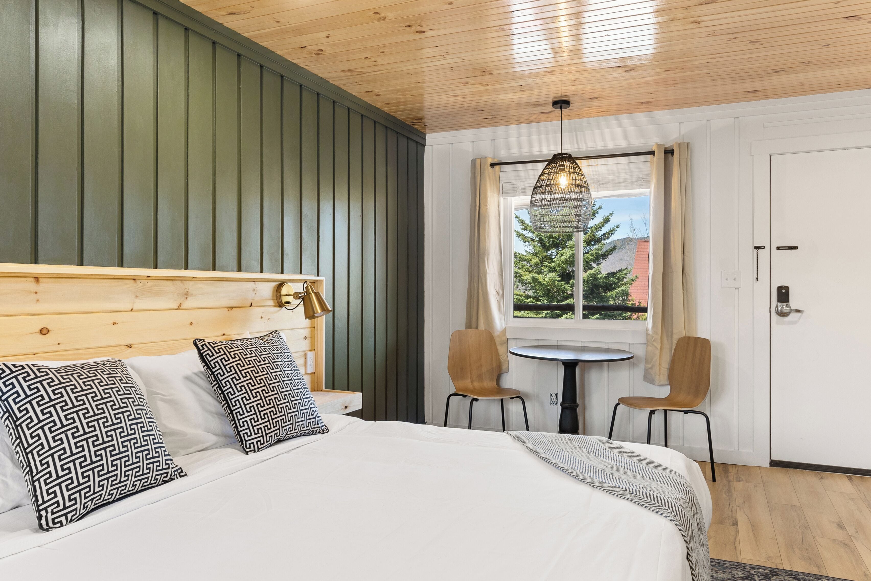 A bedroom with a wooden headboard, two patterned pillows, a bed, a hanging light fixture, a window with curtains, and two chairs.