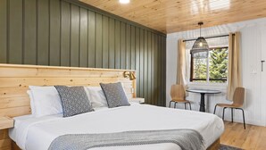 A bedroom with a wooden headboard, a large bed with patterned pillows, a small round table, two chairs, and a window with curtains. - The Boha Hotel (Lake Placid)
