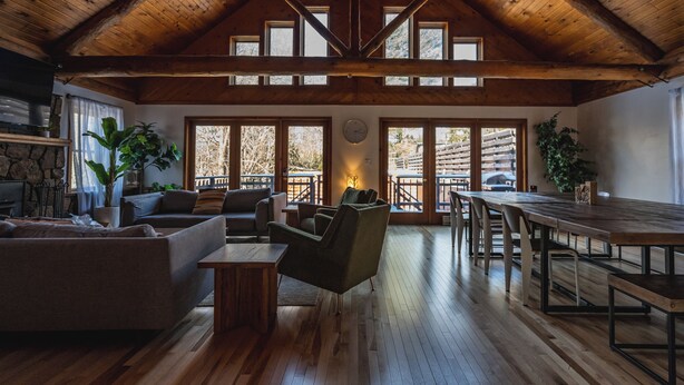 A spacious living area with wooden flooring, a high ceiling with exposed beams, a fireplace, and large windows offering views of the outdoors. - The Boha Hotel (Lake Placid)