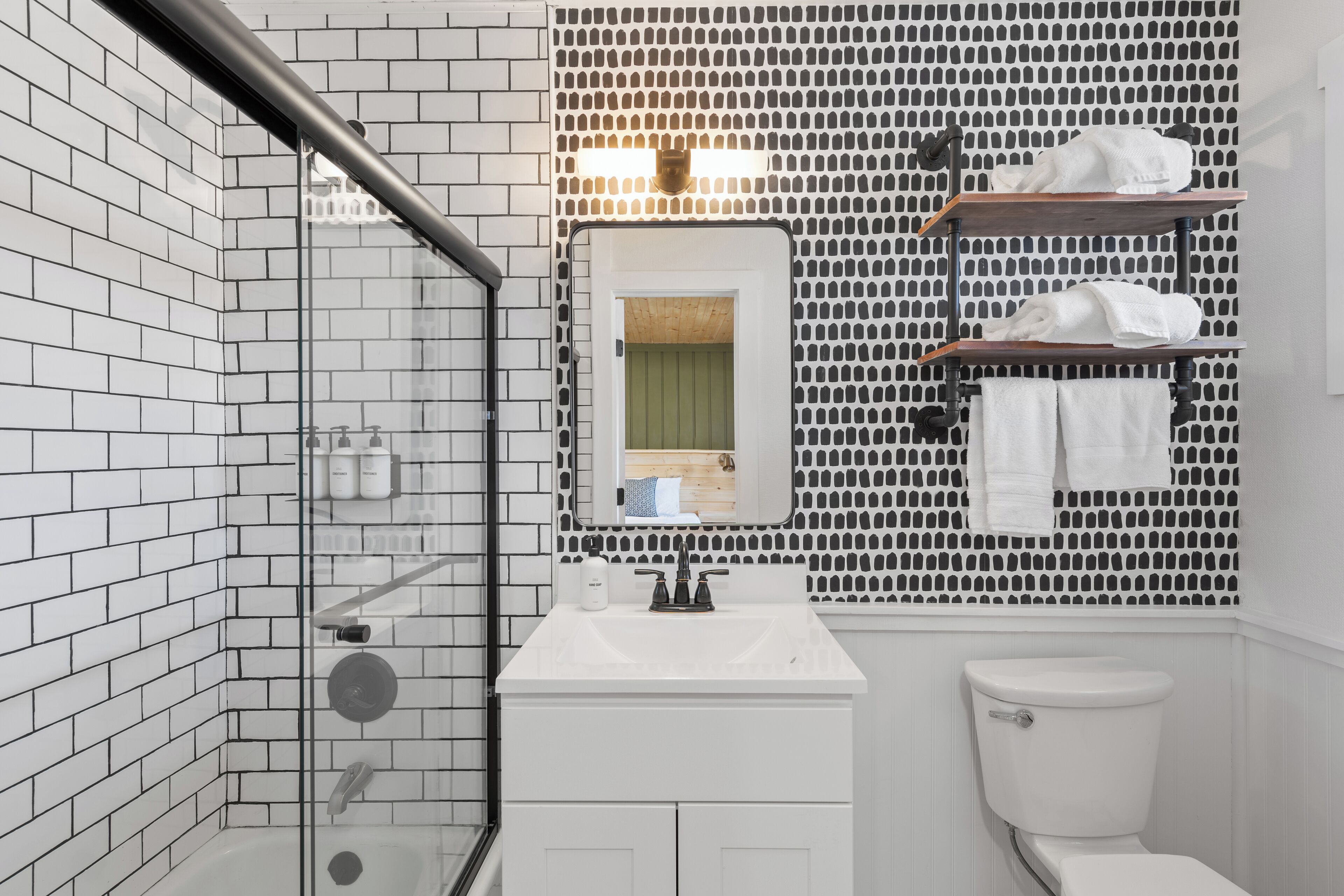A bathroom with a glass shower enclosure, white tiled walls, a white sink with a mirror, and a toilet.