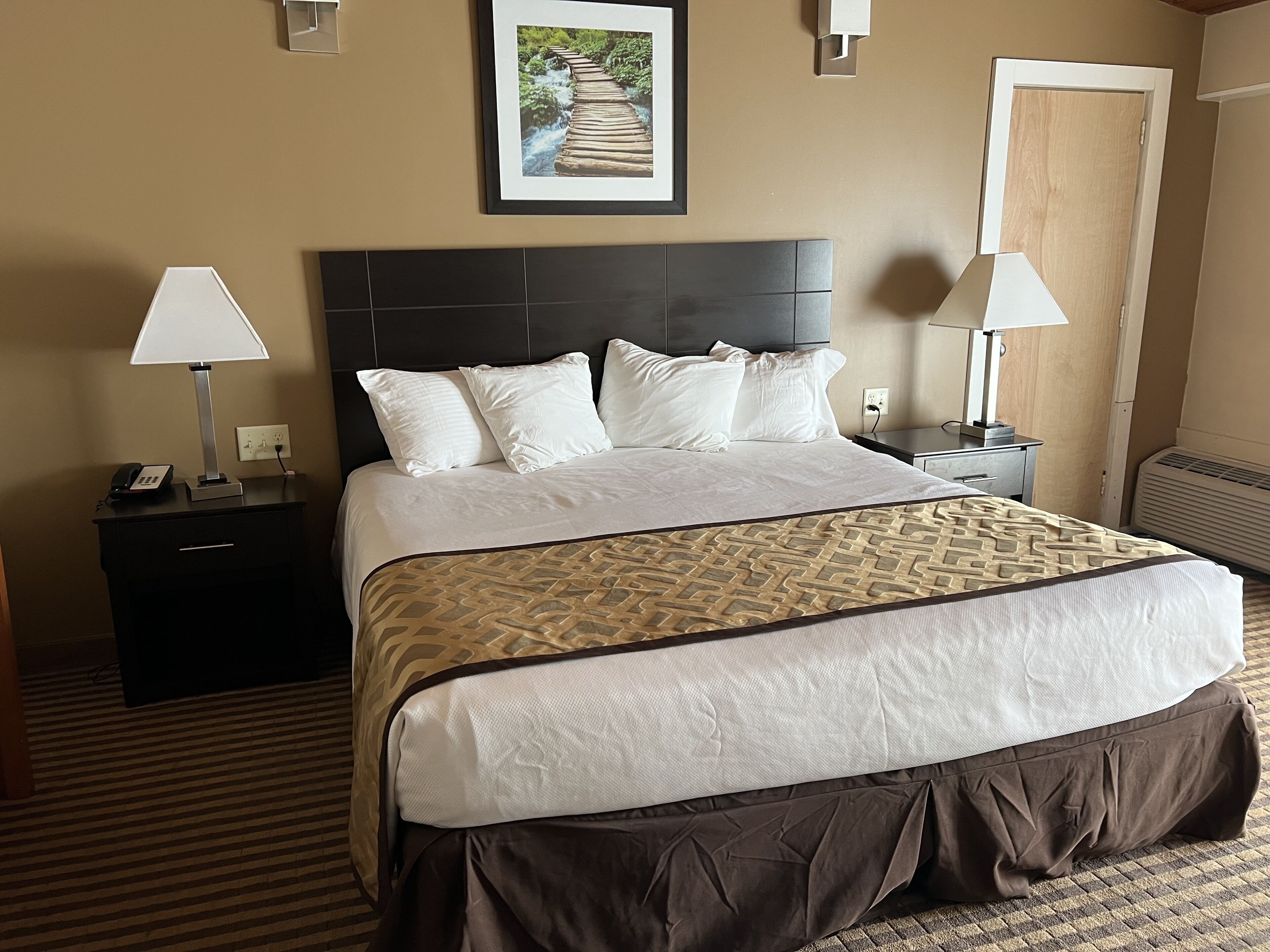 A hotel room with a large bed, two bedside tables with lamps, a nightstand with a phone, a framed picture on the wall, and a door.