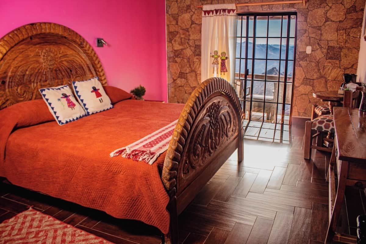 Une chambre avec un grand lit, un mur en pierre, une vue sur les montagnes par une porte vitrée et une table en bois avec des chaises.