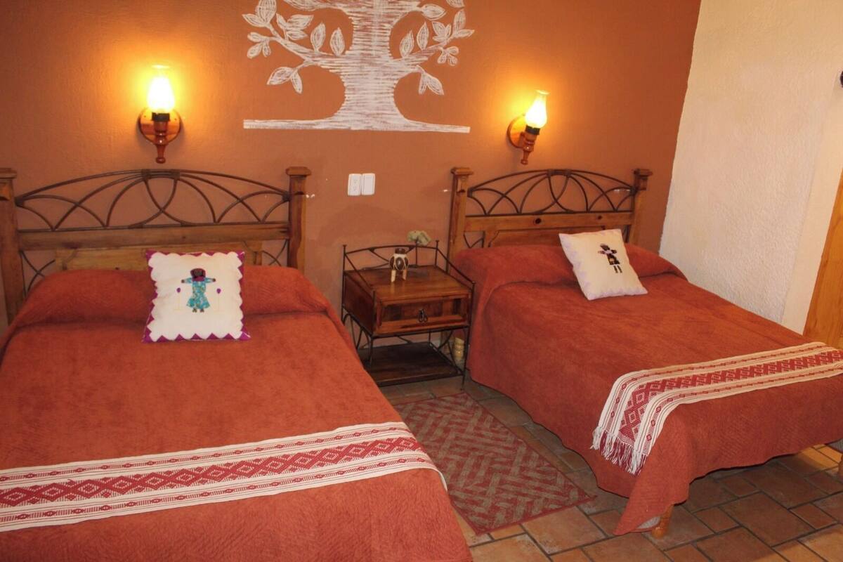 Une chambre avec deux lits, chacun recouvert d’une courtepointe rouge et agrémenté d’un coussin décoratif. De chaque côté d’une fresque murale, on trouve des lampes fixées au mur.