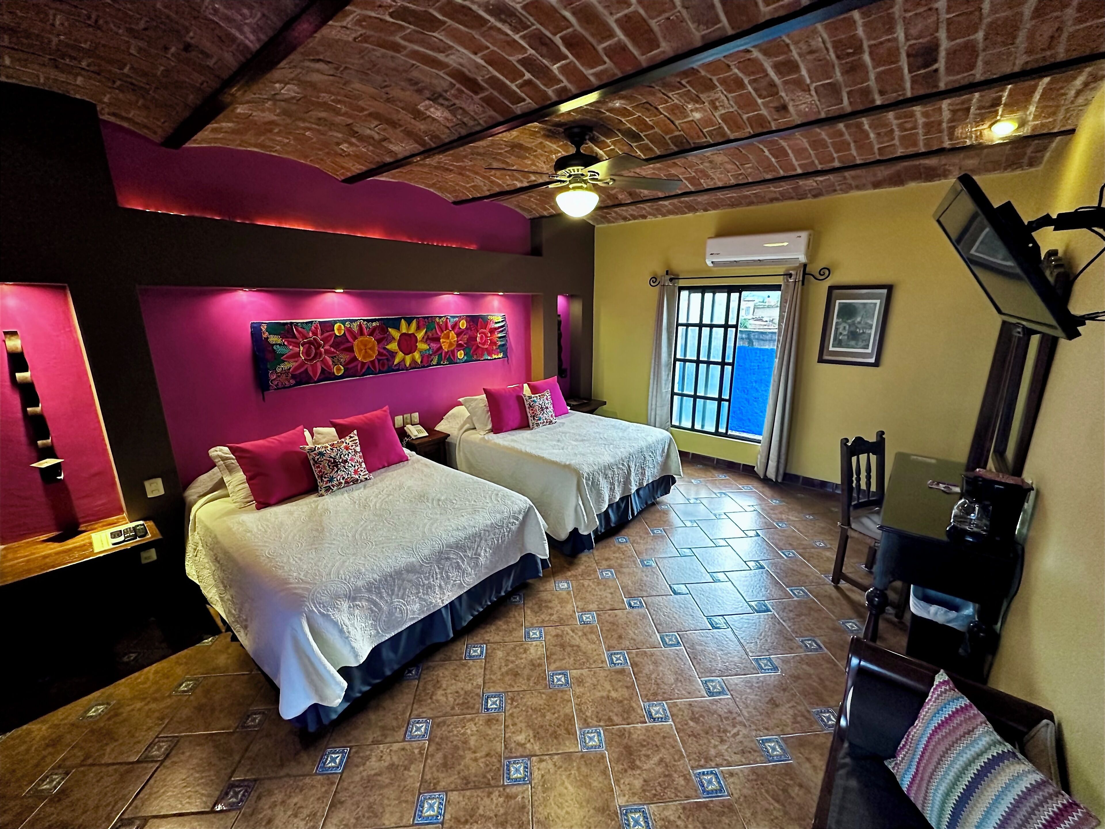 Habitación de hotel con dos camas, techo de ladrillo, un mural colorido, ventana con vistas y televisión de pantalla plana.
