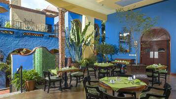An outdoor dining area with multiple tables and chairs, surrounded by potted plants and a blue wall.