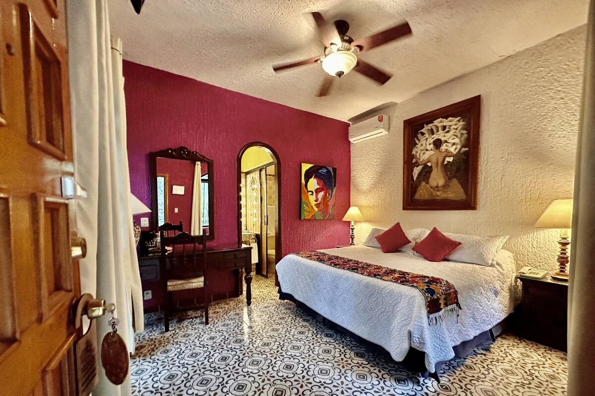 A hotel room with a bed, a desk, a chair, a mirror, a ceiling fan, and patterned floor tiles.