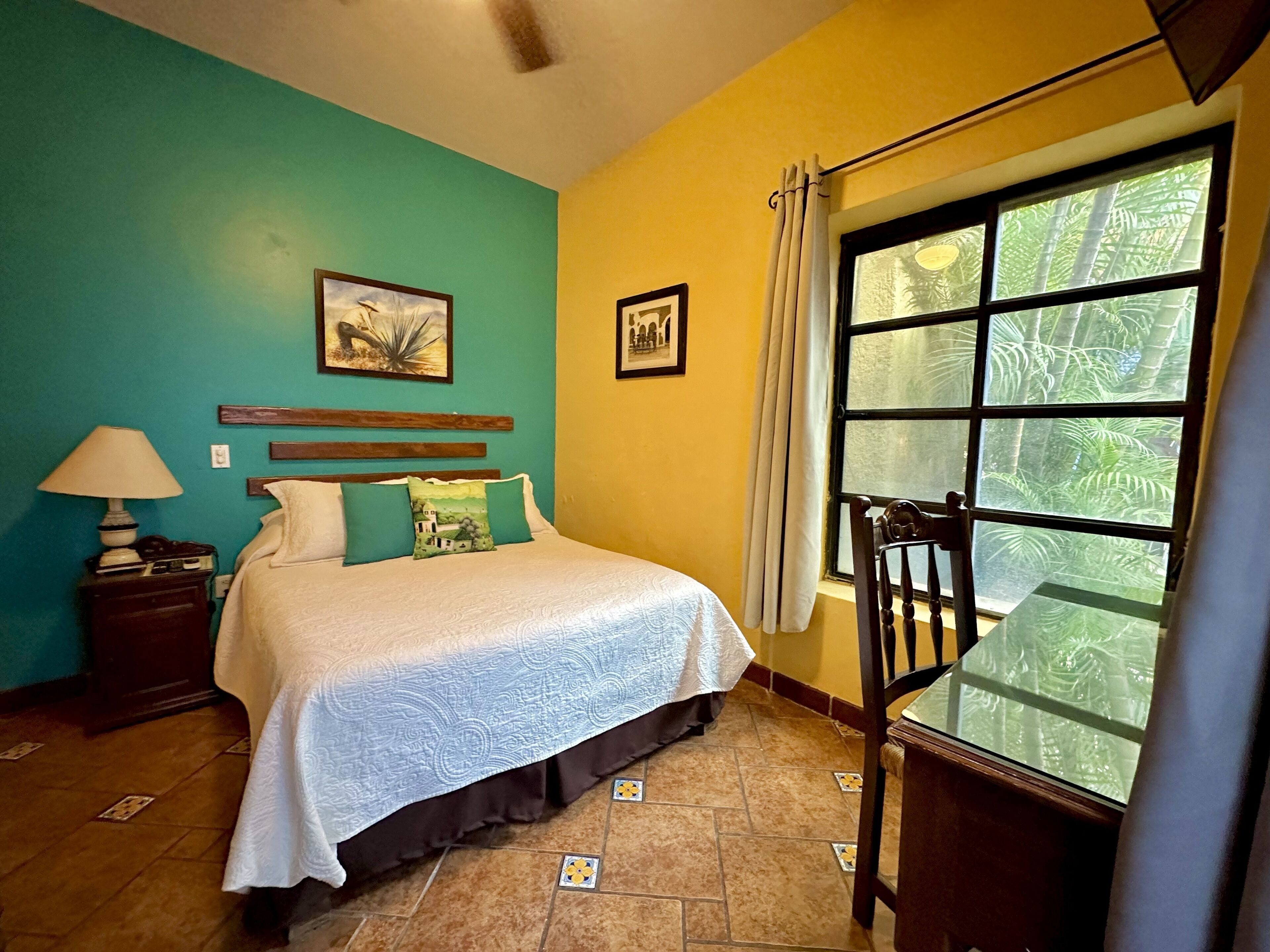 Un dormitorio con cama, mesita de noche, silla, ventana con cortinas y pared con dos cuadros.