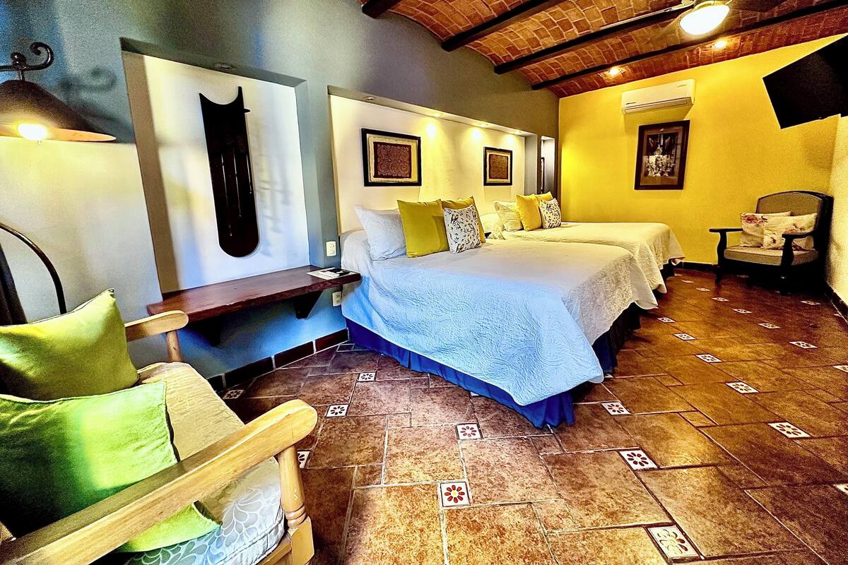 A hotel room with two beds, a wooden bench, a chair, and a tiled floor.