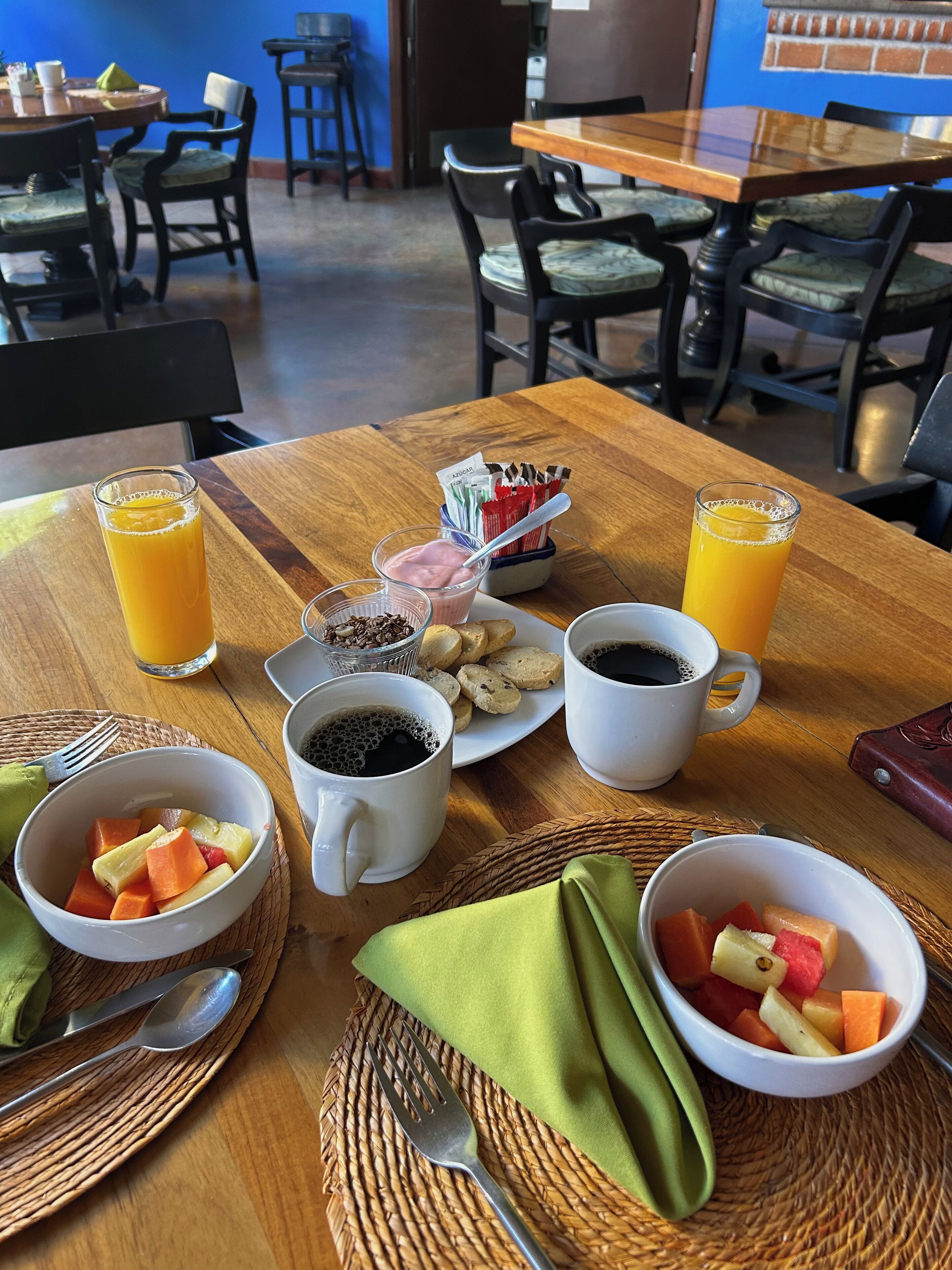 A breakfast setting with a wooden table, two glasses of orange juice, a plate of pastries, a bowl of fruit, and two cups of coffee.
