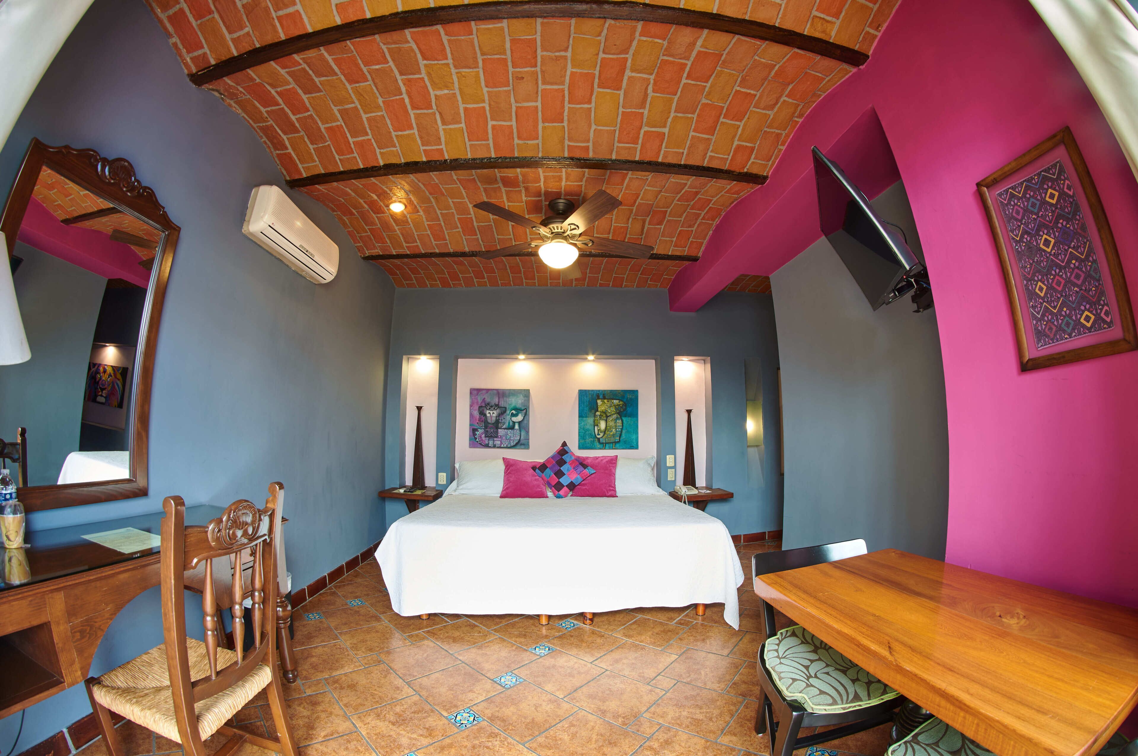 A bedroom with a brick ceiling, a bed with white bedding, a desk with a chair, and a dining table.