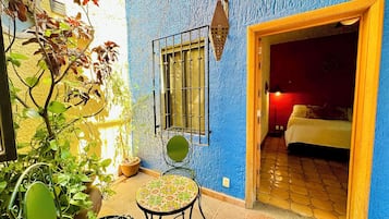 A room with a bed, a window with bars, a potted plant, and a table with a chair.