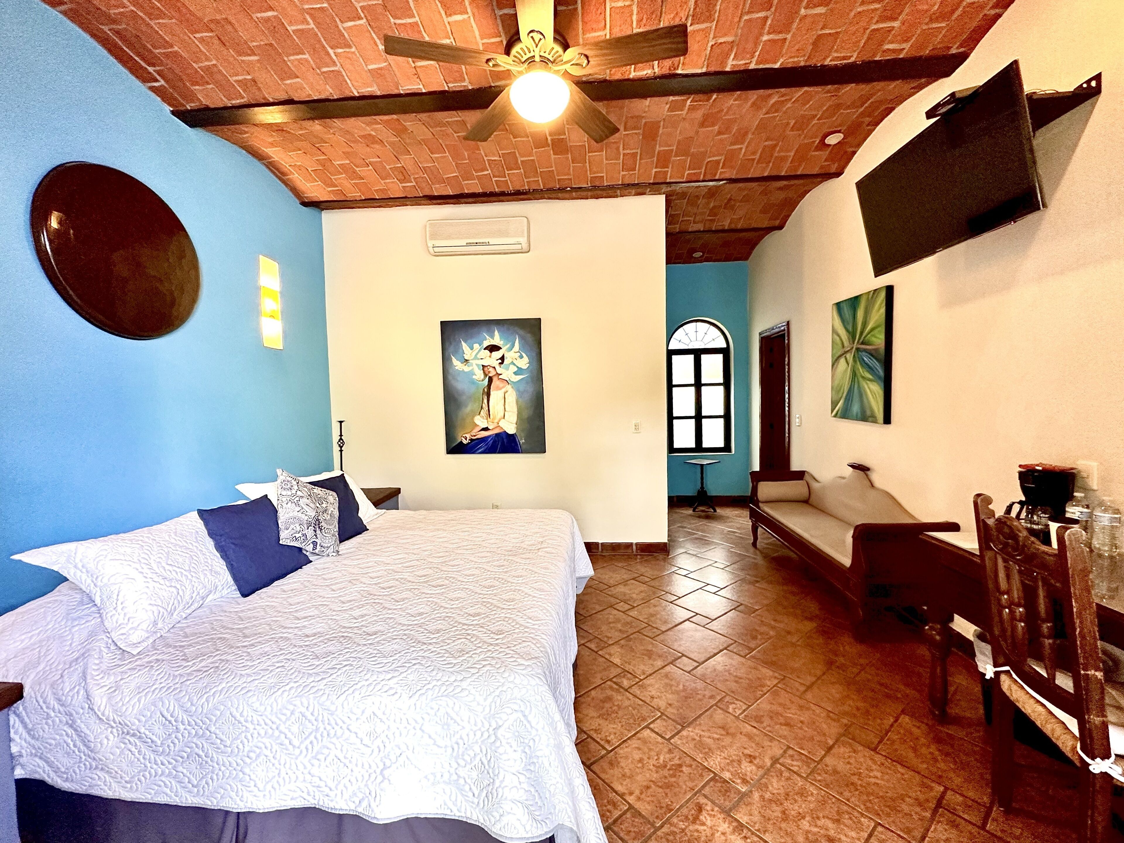 A hotel room with a bed, a ceiling fan, a painting, a window, and a TV mounted on the wall.