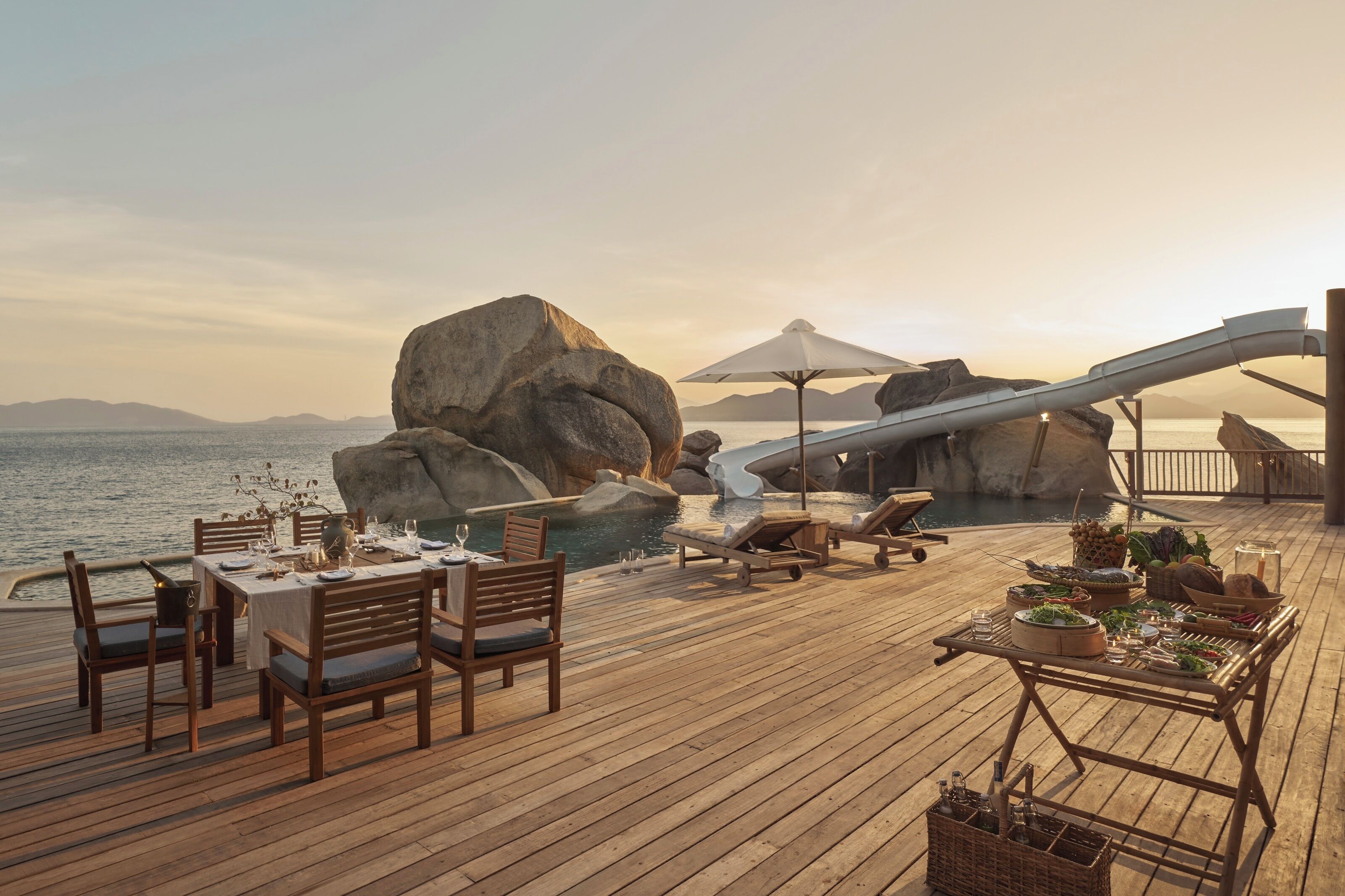 A wooden deck with a table set for dining, lounge chairs, and a pool area with a slide.