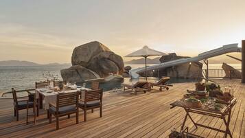 A wooden deck with a table set for dining, lounge chairs, and a pool area with a slide.