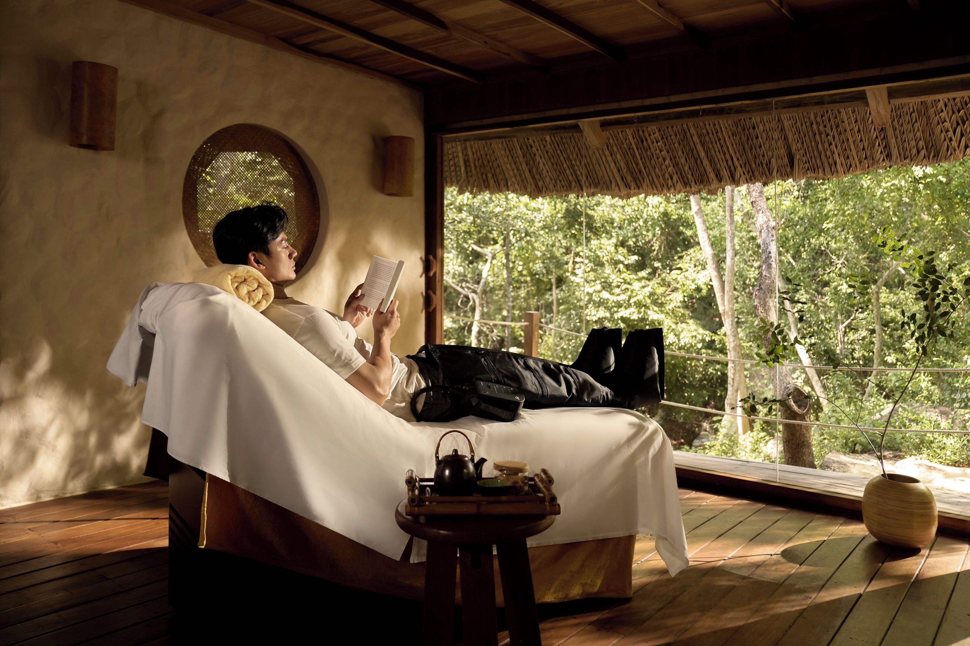 A room with a bed, a window with a view of trees, a small table with a teapot, and a wall-mounted light.