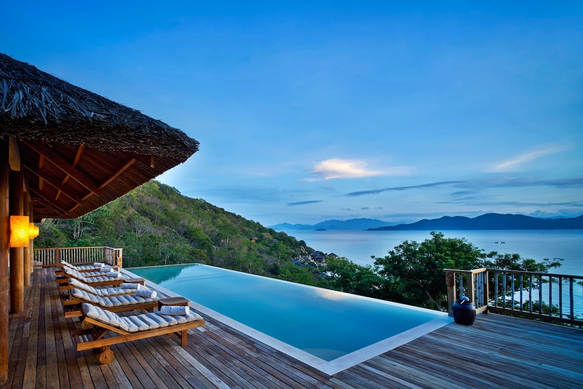 Una terraza de madera con sillones de descanso y una piscina, con vistas a un paisaje montañoso y una masa de agua.