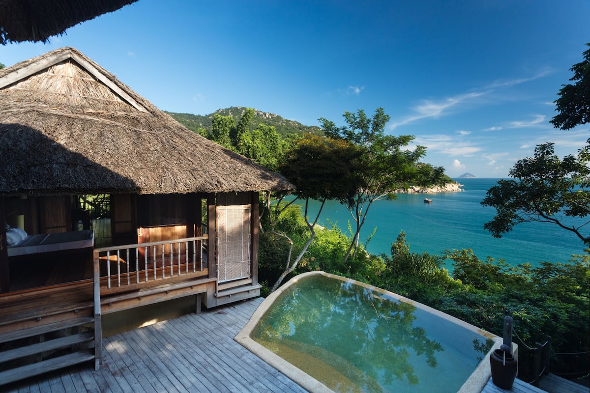 Una terraza de madera con una estructura de techo de paja, una piscina pequeña y vista al mar y una isla distante.