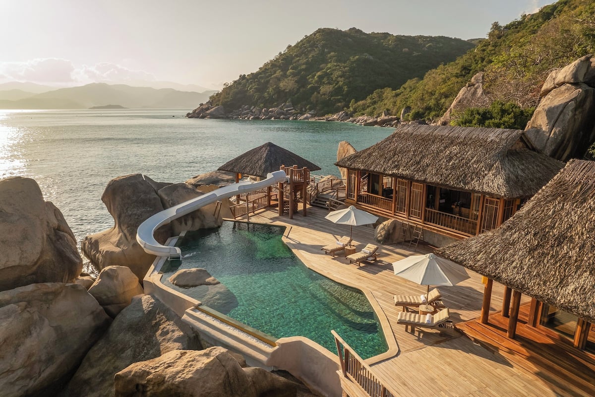Un resort con piscina cristalina, terraza de madera, cabañas con techo de paja y vistas al mar y colinas.