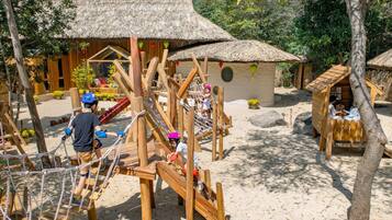 A wooden play structure with rope bridges and slides, surrounded by thatched-roof huts and trees.