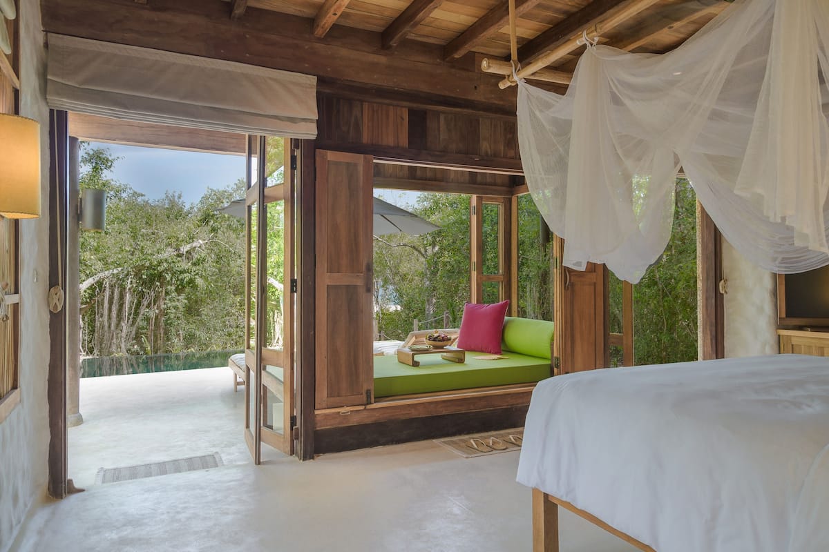 Un dormitorio con cama, un sofá verde con cojines, techo de madera y vistas a árboles a través de amplias ventanas.