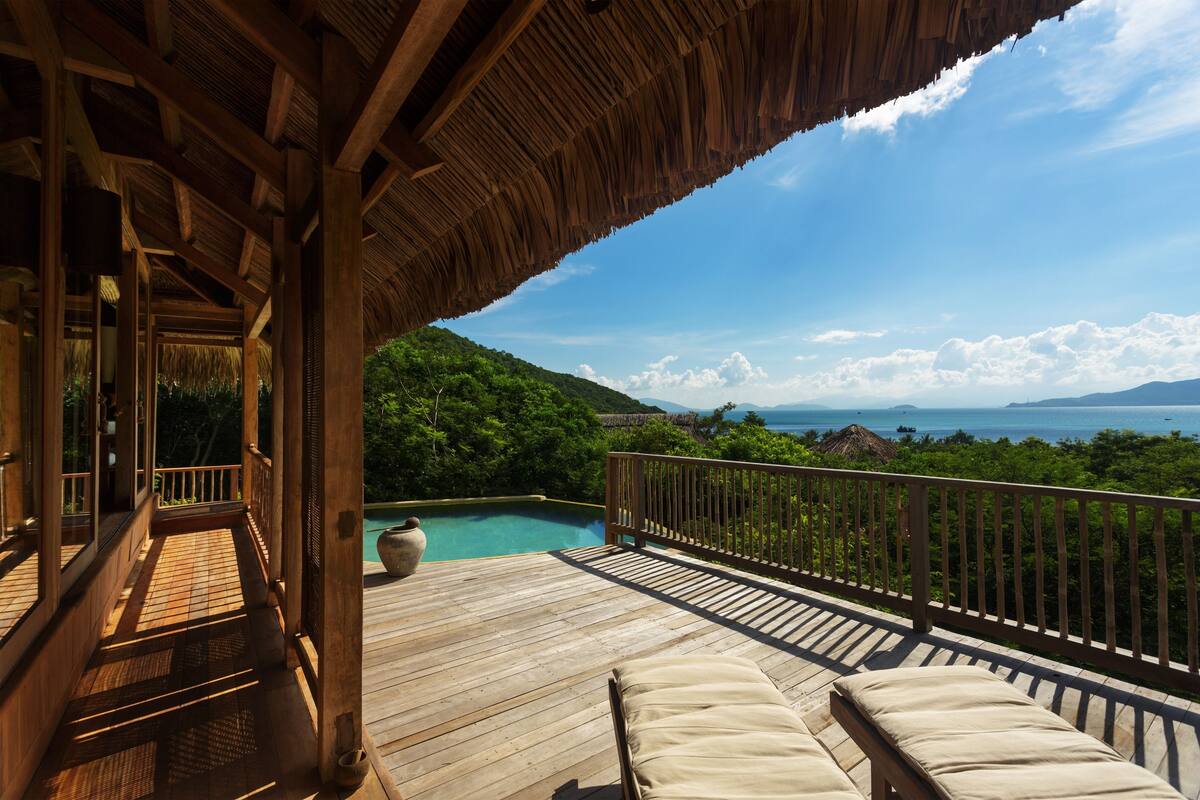 Una terraza de madera con techo de paja que da a una piscina y una vista panorámica de un paisaje exuberante y el océano.
