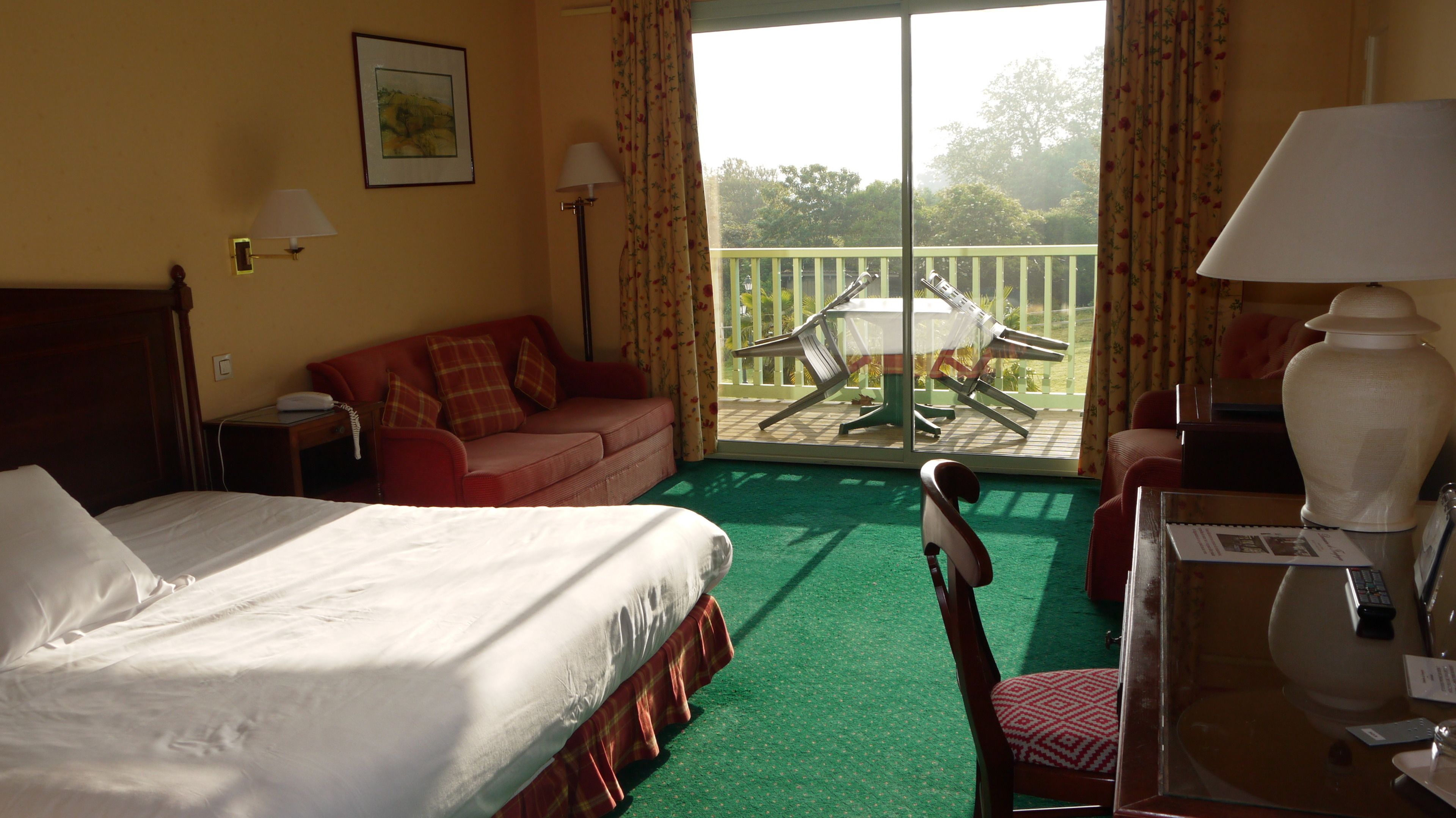 Une chambre d’hôtel avec un lit, un canapé rouge, un balcon avec une table et des chaises, et une vue sur l’extérieur.