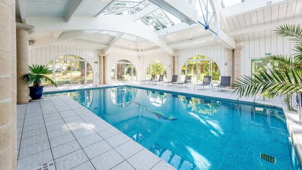 An indoor swimming pool with clear blue water, surrounded by white tiled flooring and columns.
