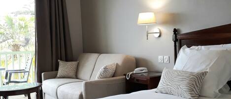 Una habitación de hotel con una cama, dos sillones, una mesita pequeña y una mesita de noche con lámpara.
