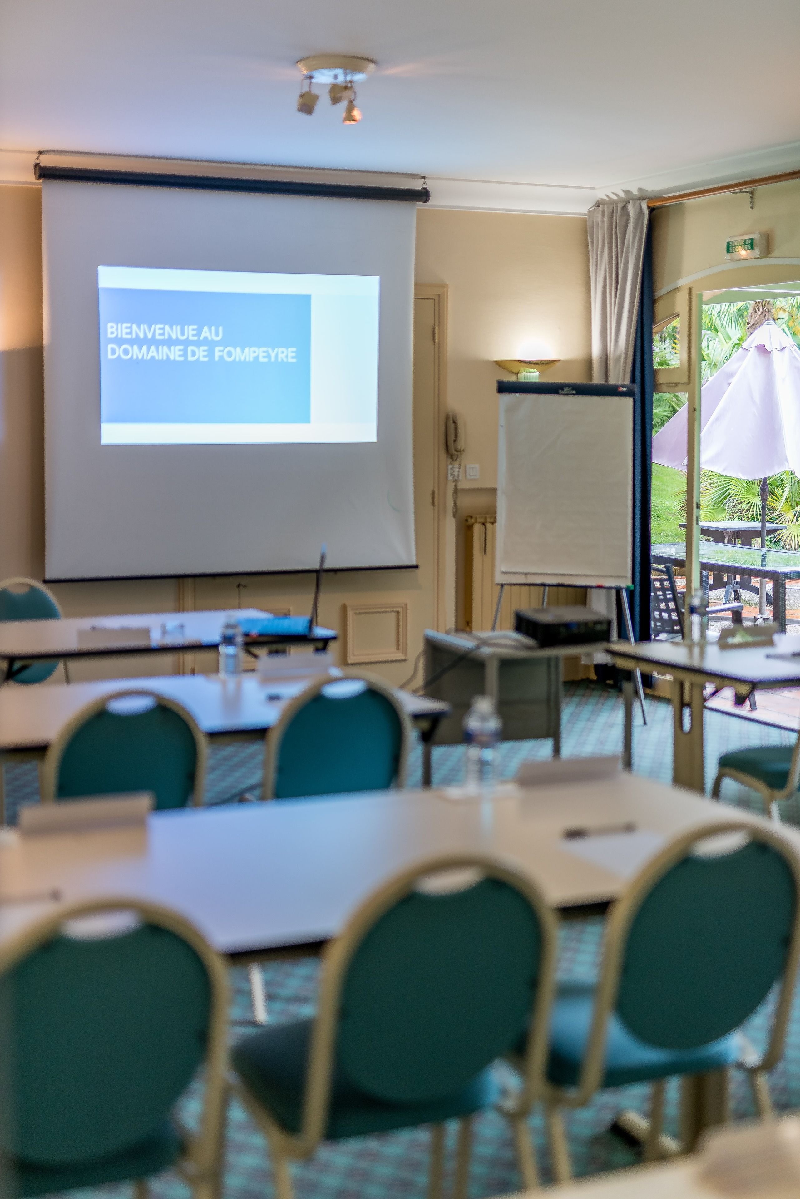 A conference room with a projection screen displaying "BIENVENUE AU DOMAINE DE FOMPEYRE," tables, chairs, a whiteboard, and a view of the outdoors.