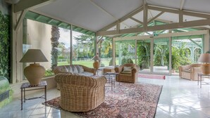 A spacious indoor area with wicker furniture, a patterned rug, and large windows offering a view of greenery outside.
