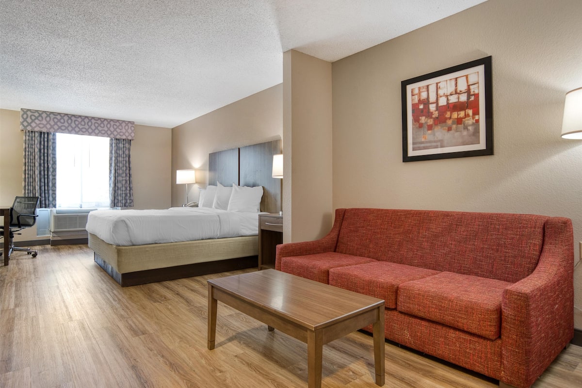 Ein Hotelzimmer mit einem Bett, einem roten Sofa, einem Holztisch, einem Schreibtisch mit Stuhl und einem Fenster mit Vorhängen.