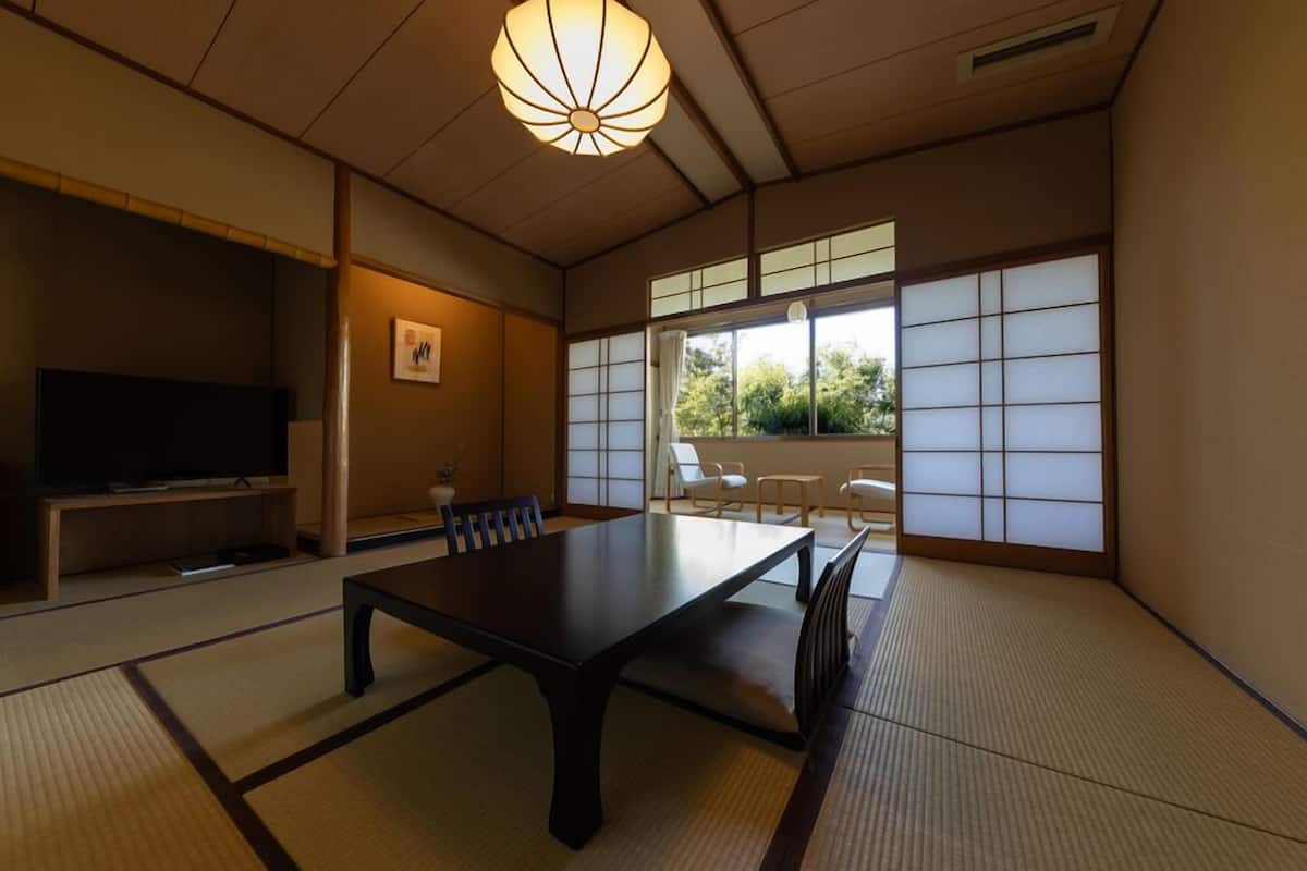 Una habitación japonesa tradicional con suelo de tatami, una mesa baja, sillas, televisor y una puerta corrediza con vistas.