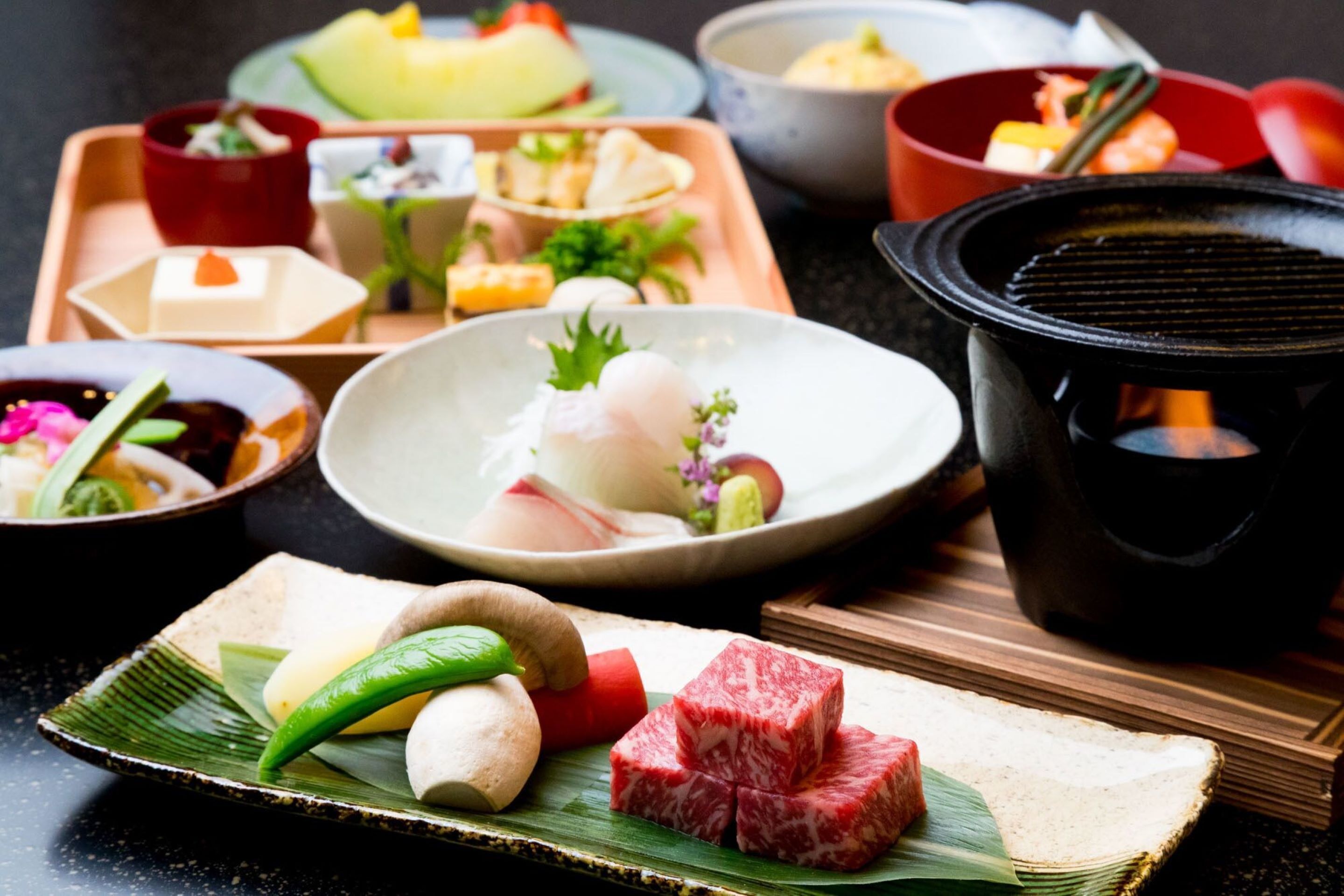 A variety of Japanese dishes on a wooden tray, including sushi, sashimi, and other delicacies, with a black pot in the background.