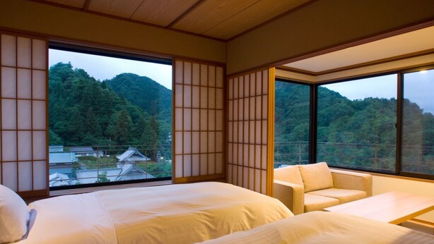 Un dormitorio tradicional japonés con suelo de tatami, dos camas, un sofá y grandes ventanales con vistas a una montaña boscosa.