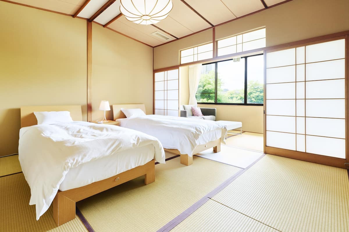 Une pièce de style japonais traditionnel, avec un sol recouvert de tatamis, deux lits en bois avec du linge de lit blanc et une grande fenêtre donnant sur un paysage verdoyant.