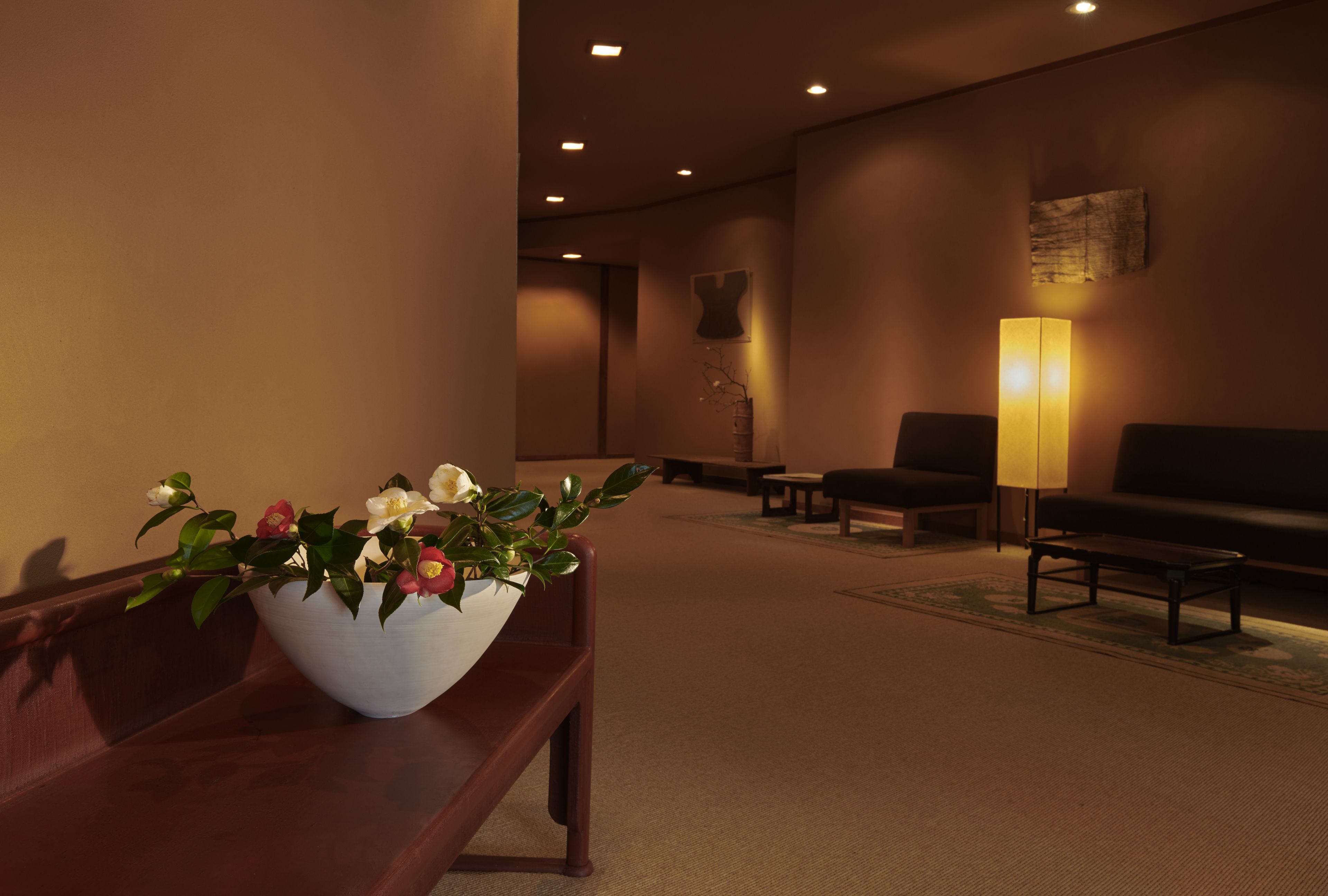 A lobby with a wooden bench, a white vase with flowers, a sofa, a coffee table, and a floor lamp.